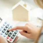 Une femme fait un calcul avec une calculatrice devant une maison miniature.